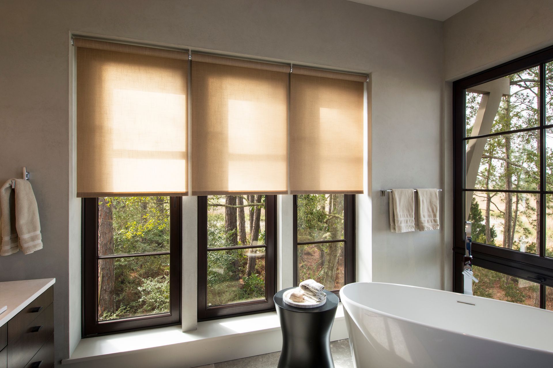 Bathroom with window and beige roller shades, a soaking tub, and a view of trees.