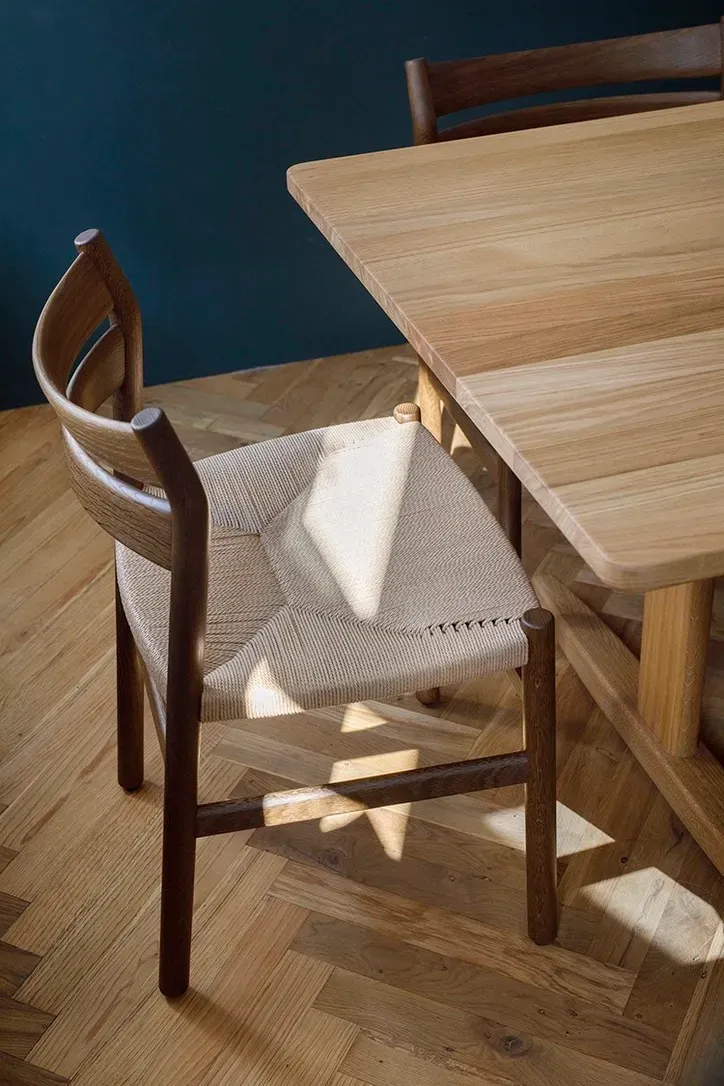 Wooden chair with woven seat next to a wooden table, set on a wood floor. Sunlight casts shadows.