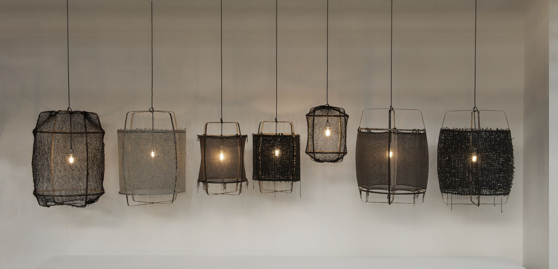 Seven pendant lights of varying shapes and designs, each with a lit bulb, hanging against a neutral wall.