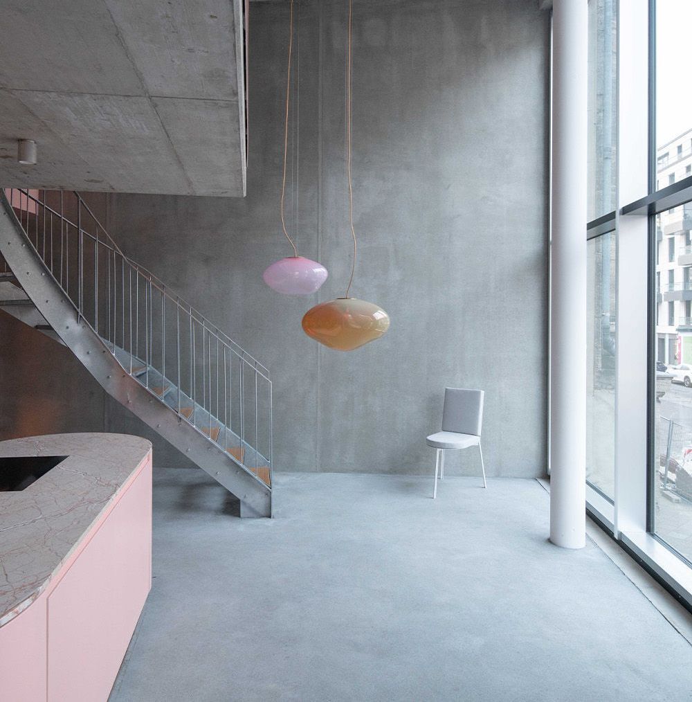 Modern interior with a concrete staircase, pink countertop, and two hanging spherical lights. A chair sits against a gray wall, next to a large window.