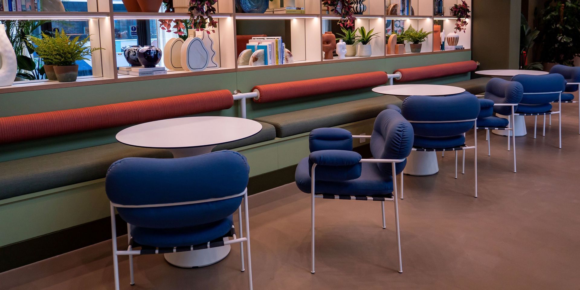 A modern café with blue chairs, white tables, and a green bench with orange bolsters. Shelves display decorative items.