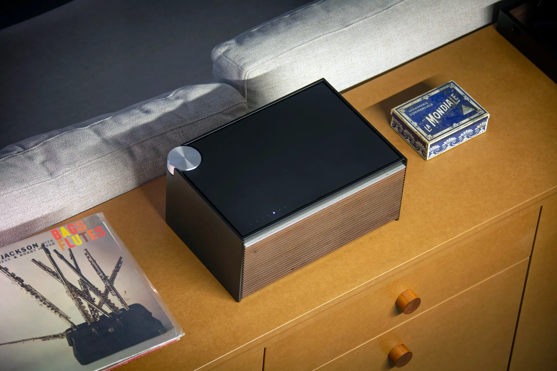 A stylish speaker with a black top and brown mesh sides on a wooden surface, beside a magazine and decorative box.