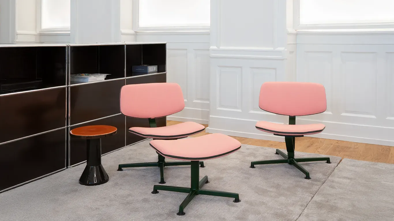 Two pink chairs and a matching footstool with dark green bases on a gray rug in a room with a black cabinet and a small side table.