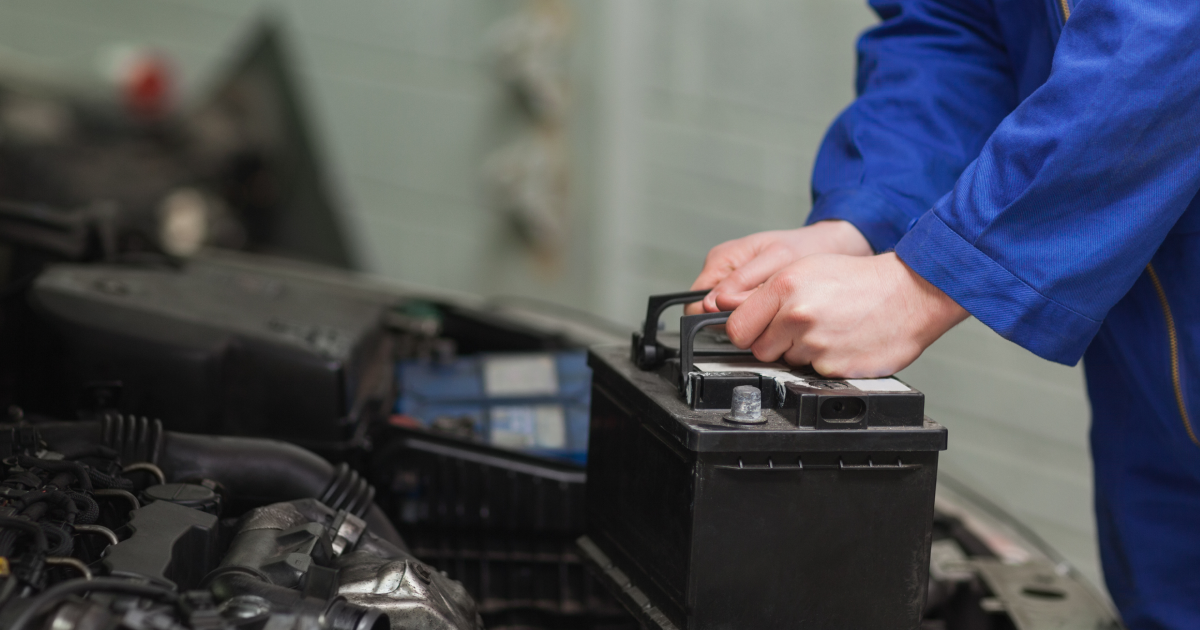 A technician in blue coveralls replaces a car battery in a workshop setting.
