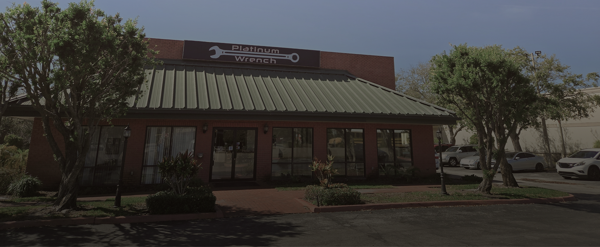 A one-story brick building with a green metal roof, a wrench logo sign, and parked cars in front on a sunny day.