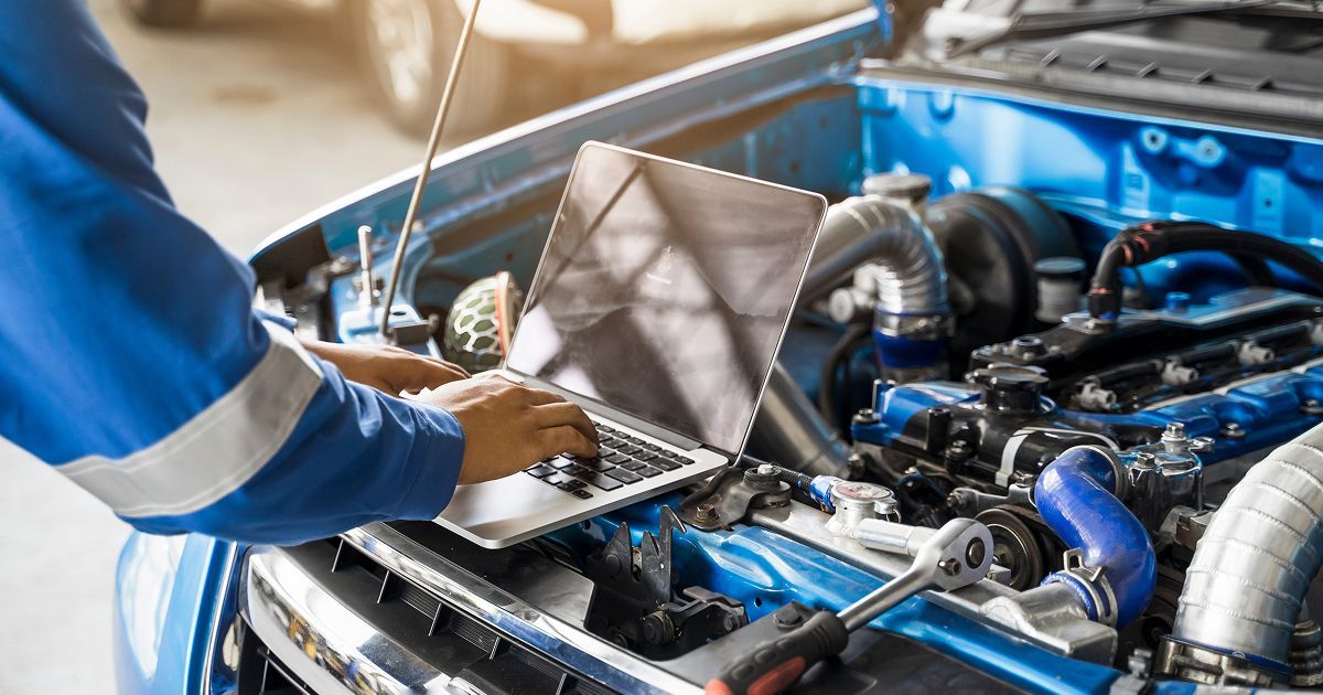 A technician in a blue uniform uses a laptop to diagnose a blue car engine under an open hood in a garage.