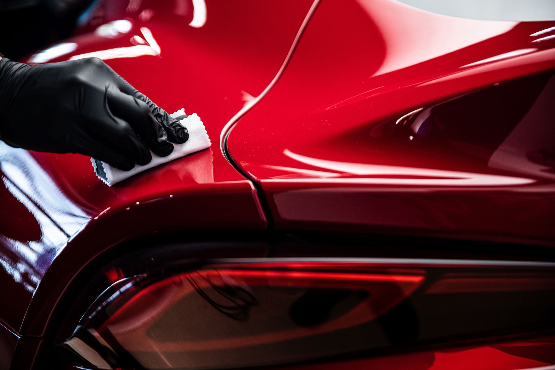A gloved hand uses a white cloth to wipe or polish the glossy red exterior of a vehicle near a tail light.
