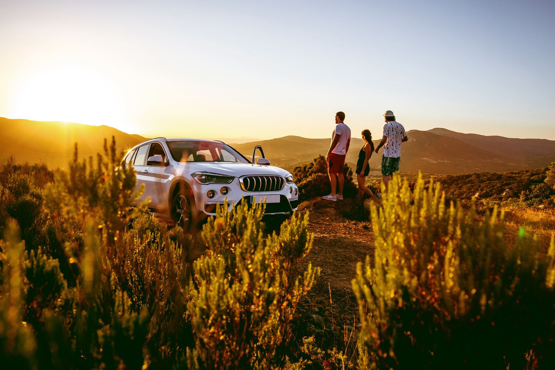 A white SUV parked on a grassy hill during sunset with three people standing nearby looking out at the mountains.