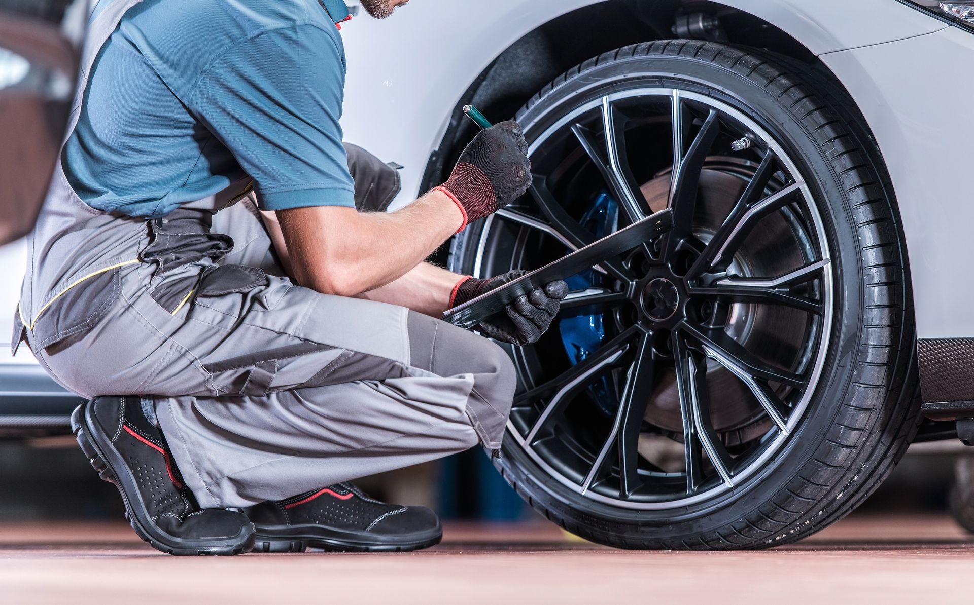 A mechanic in grey workwear squats to service the tire and wheel of a white car in a garage.