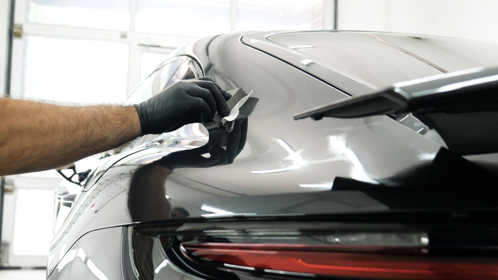A person in black gloves applies protective film to the rear panel of a silver sports car.