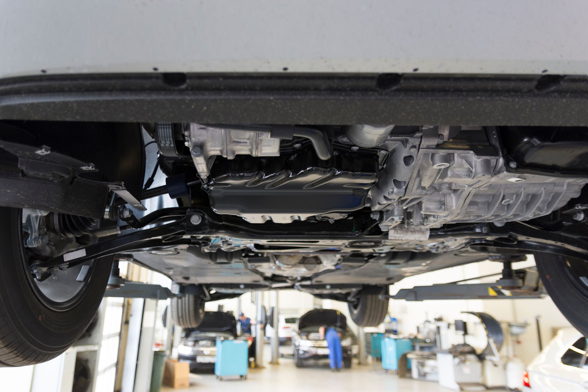 Undercarriage view of a vehicle lifted on a hydraulic hoist inside a brightly lit automotive repair shop.