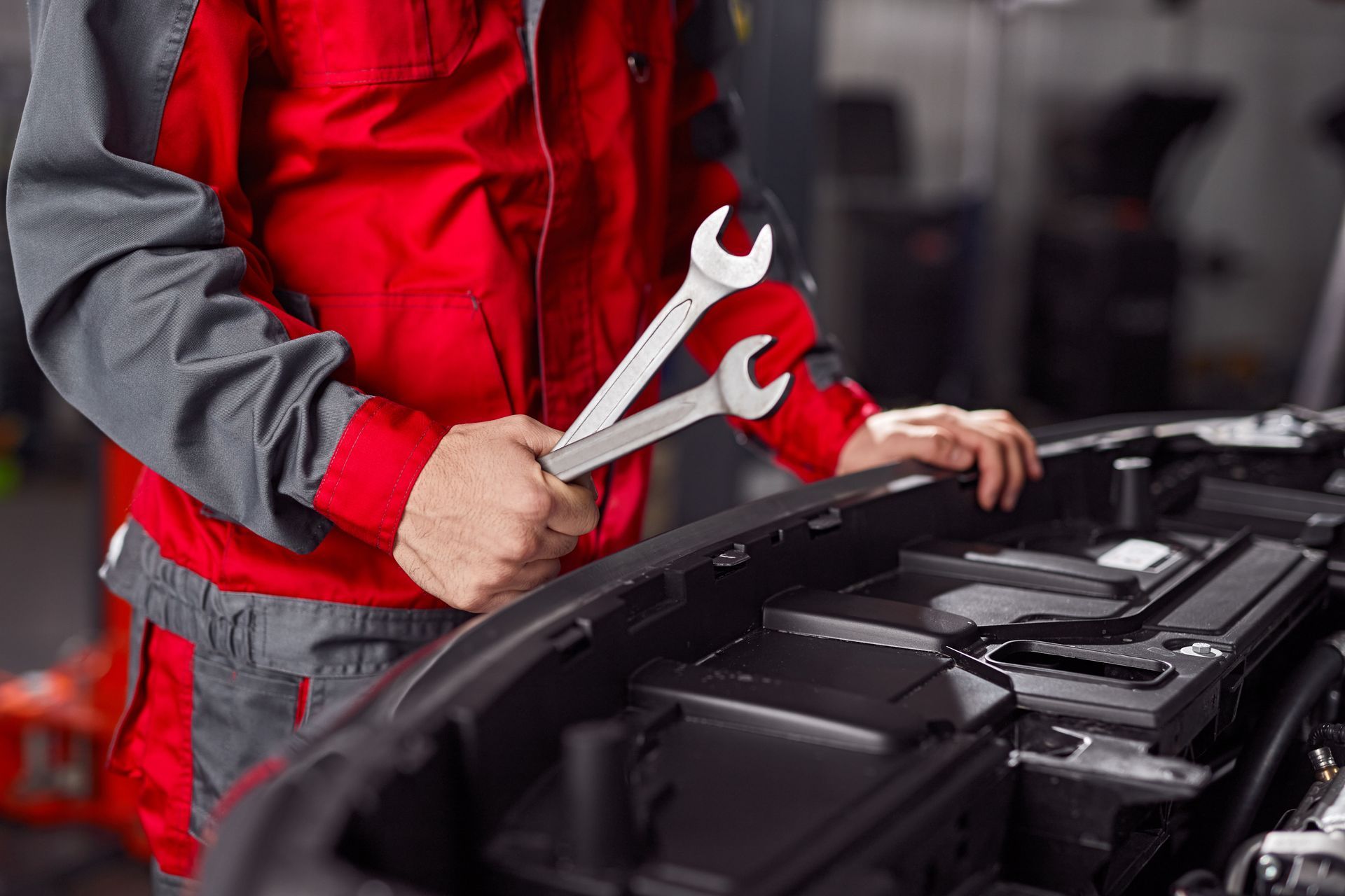 A mechanic in a red and gray uniform holds two wrenches while working on an open car engine in a garage.