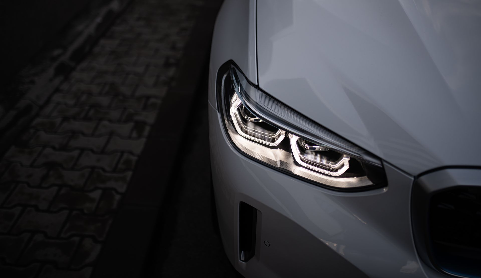 A close-up view of the illuminated, angular LED headlight and front body panel of a light-gray BMW vehicle.