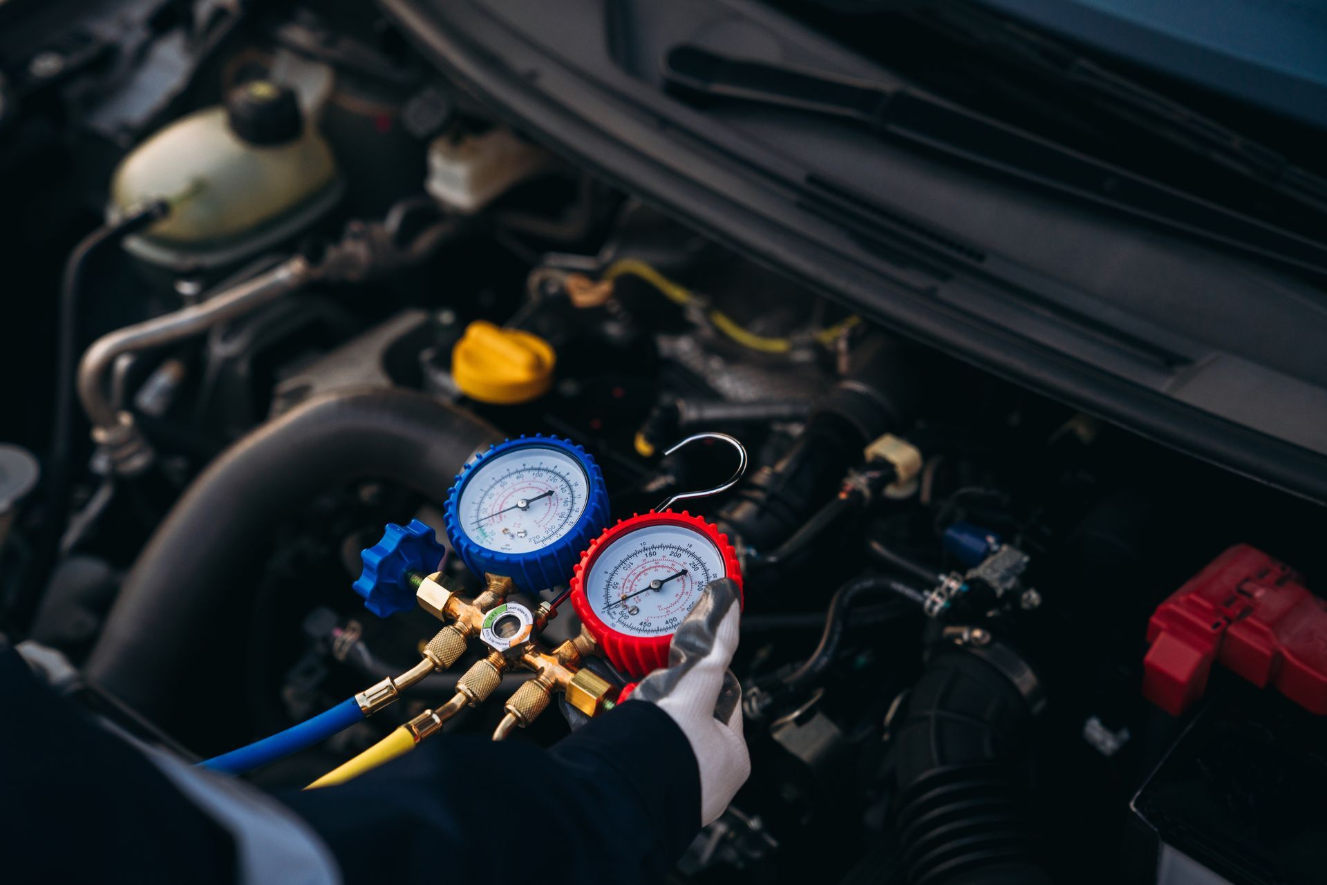 A technician in gloves checks car air conditioning levels using a red and blue gauge manifold set under the hood.