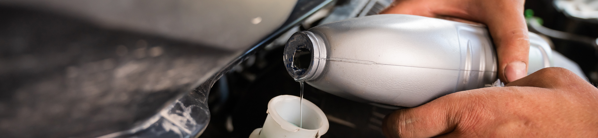 Hands pouring fluid from a plastic bottle into a car's reservoir.