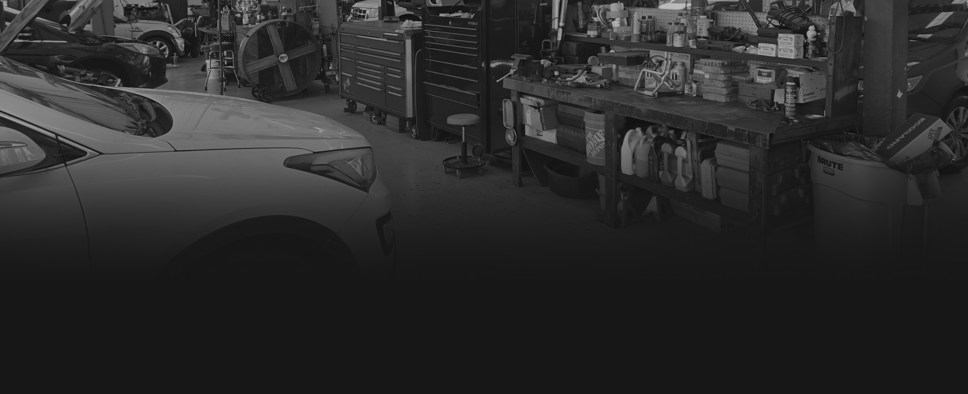 A black and white view of an automotive repair shop featuring a car in the foreground and workbenches with supplies.
