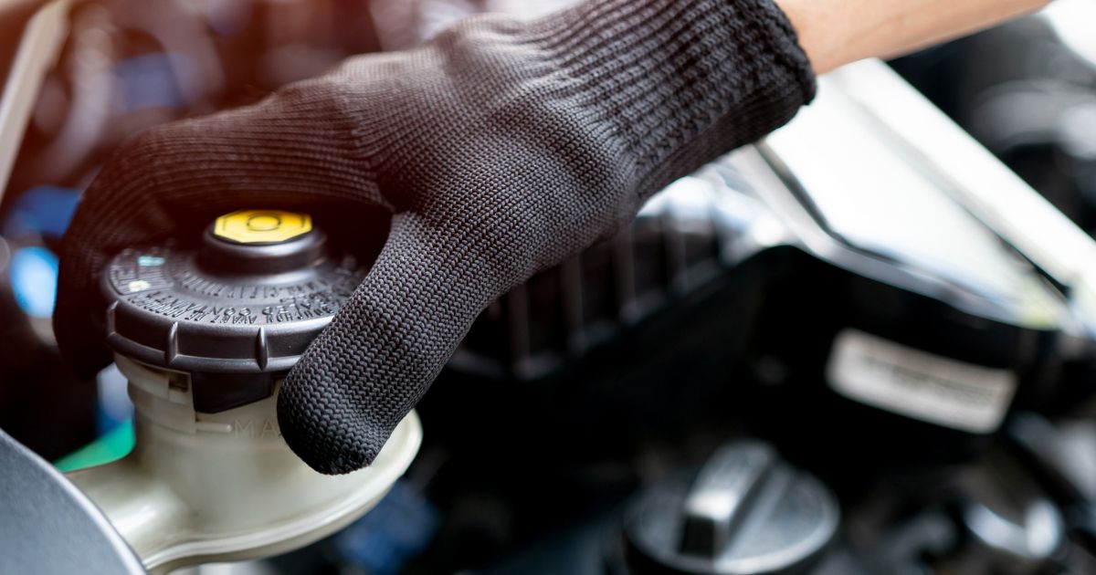 A gloved hand unscrewing the yellow-capped reservoir lid under a vehicle's hood.