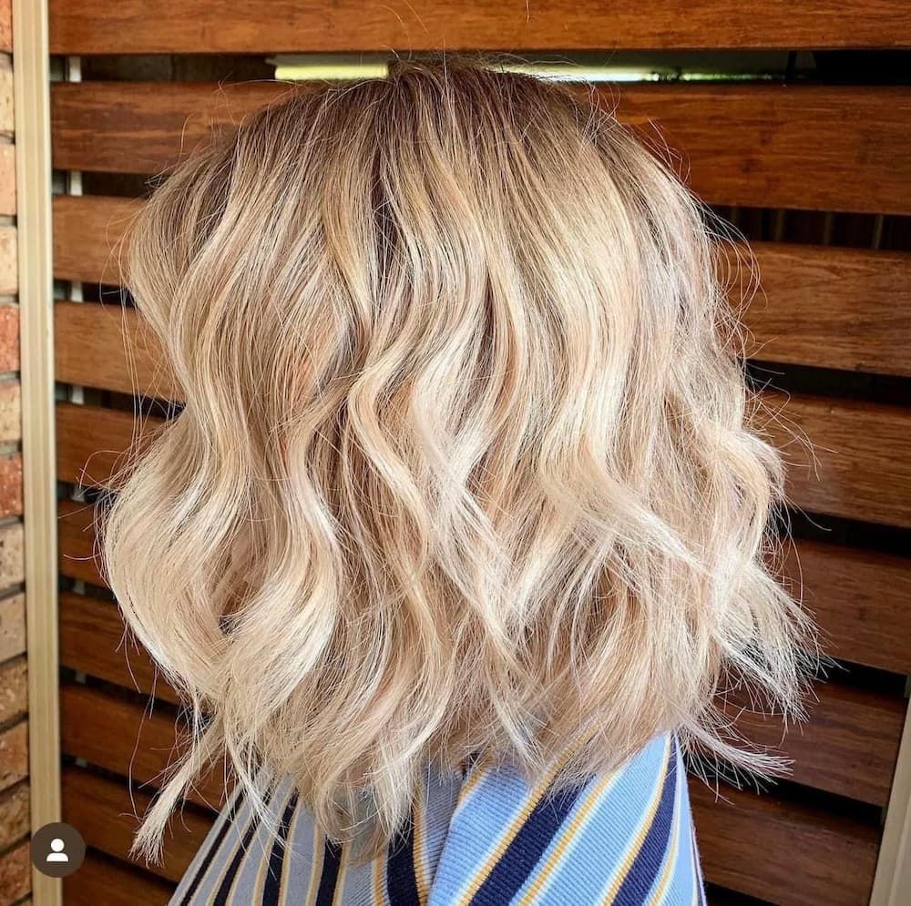 A Woman with Blonde Wavy Hair Is Standing in Front of A Wooden Fence — Apricate & Wild Salon in Airlie Beach, QLD