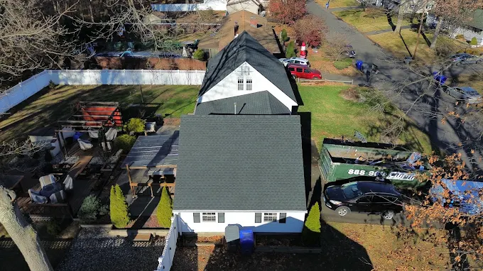 Aerial view of a white house with a dark gray roof, surrounded by a yard with trees and a car parked on the street.