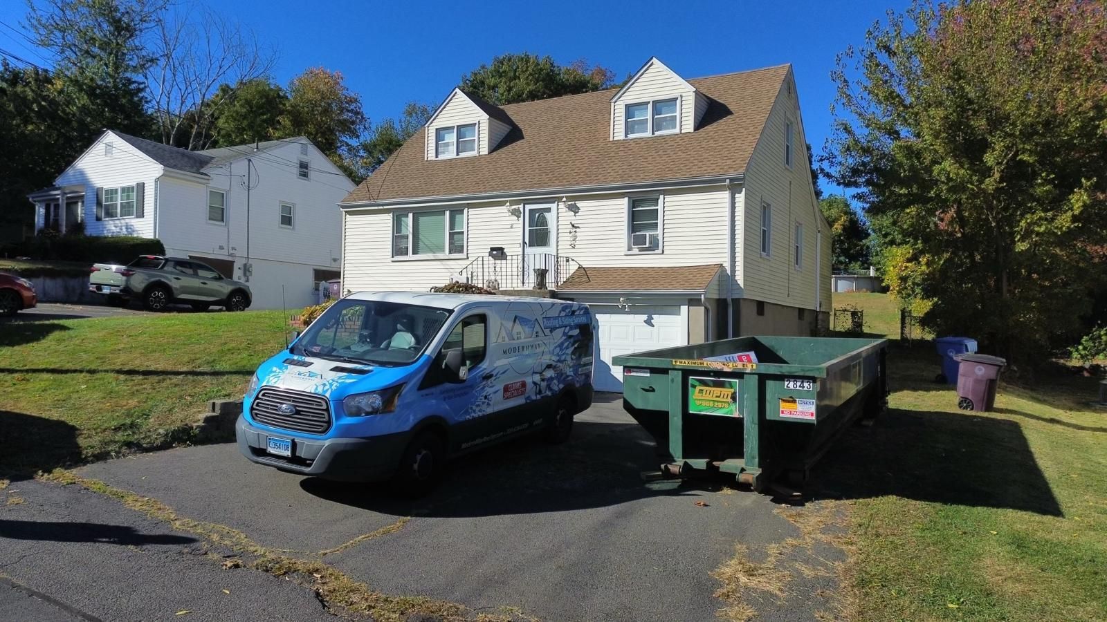 A white two-story house with a blue van and a dumpster parked in the driveway.