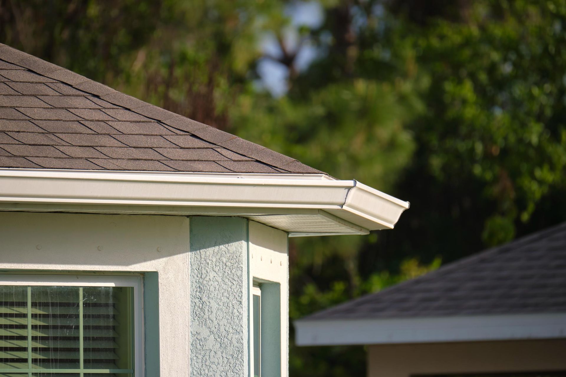 Corner of a house with brown shingled roof, white gutters, and light blue siding; trees in background.