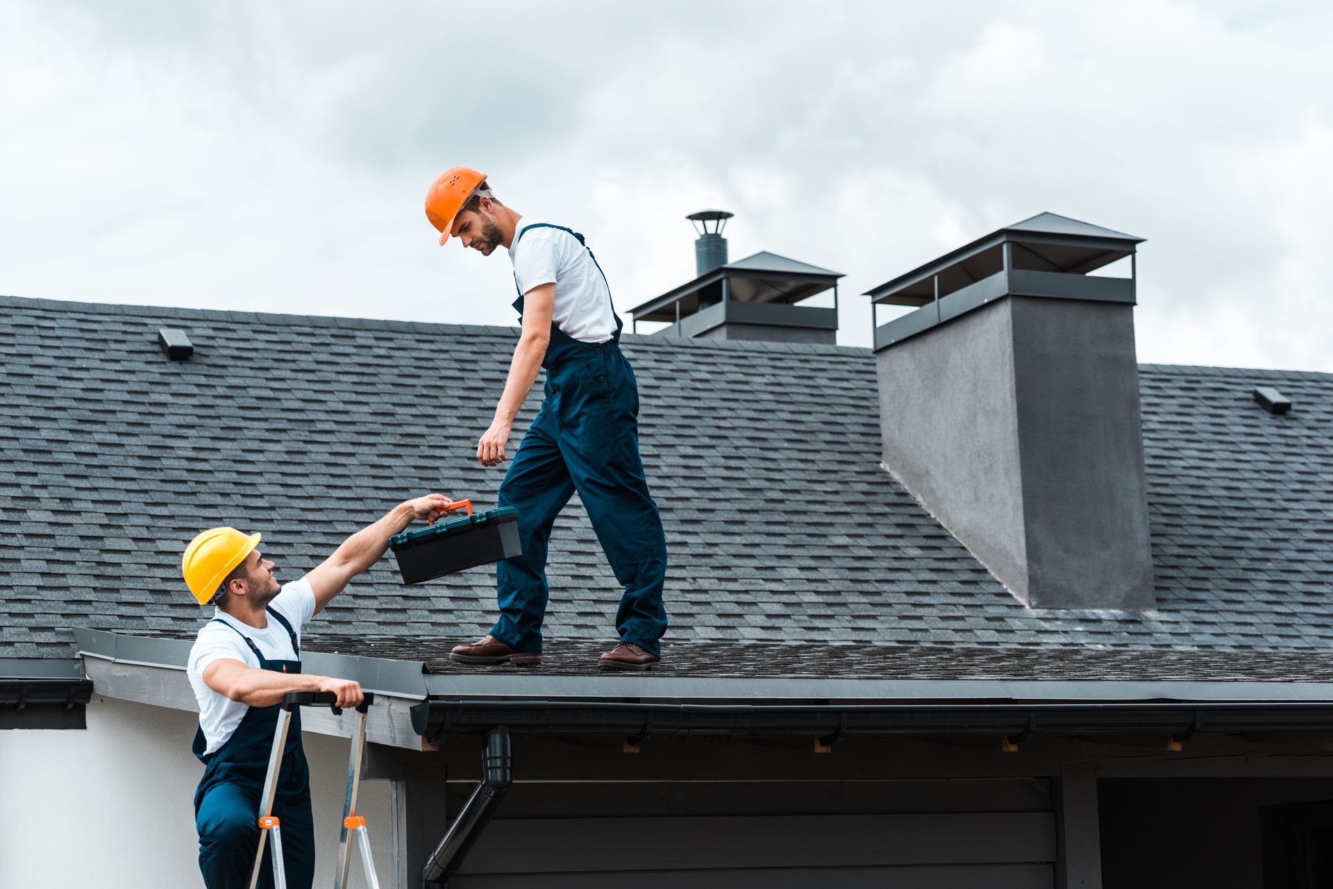 Two roofers on a rooftop, one handing a toolbox to the other.