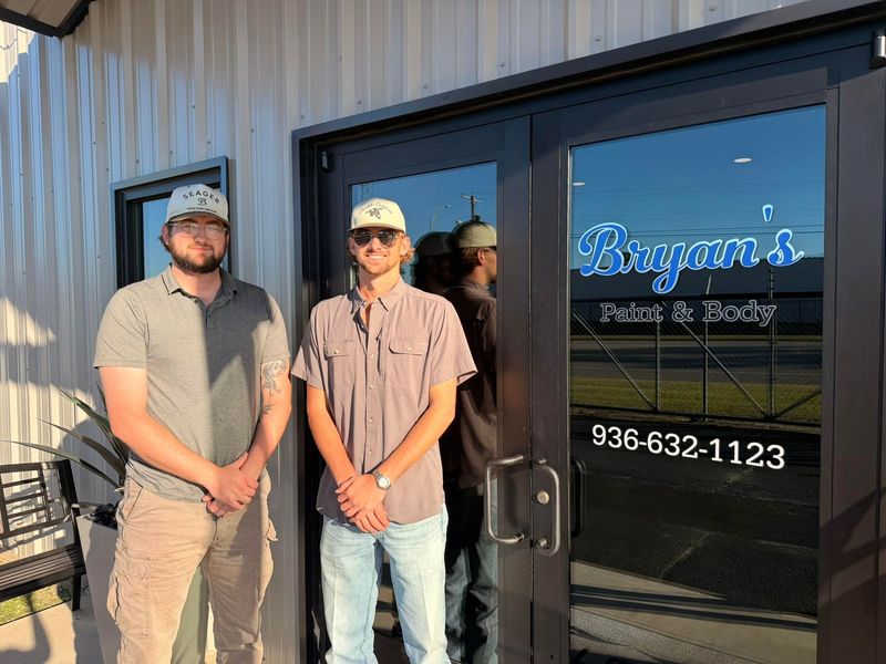 Two men in front of Bryan's Paint & Body shop. One in gray shirt and khakis, the other in a button-up shirt and jeans.