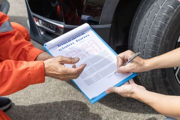 Person signing insurance claim form next to a car tire. Orange jumpsuit, clipboard, asphalt background.