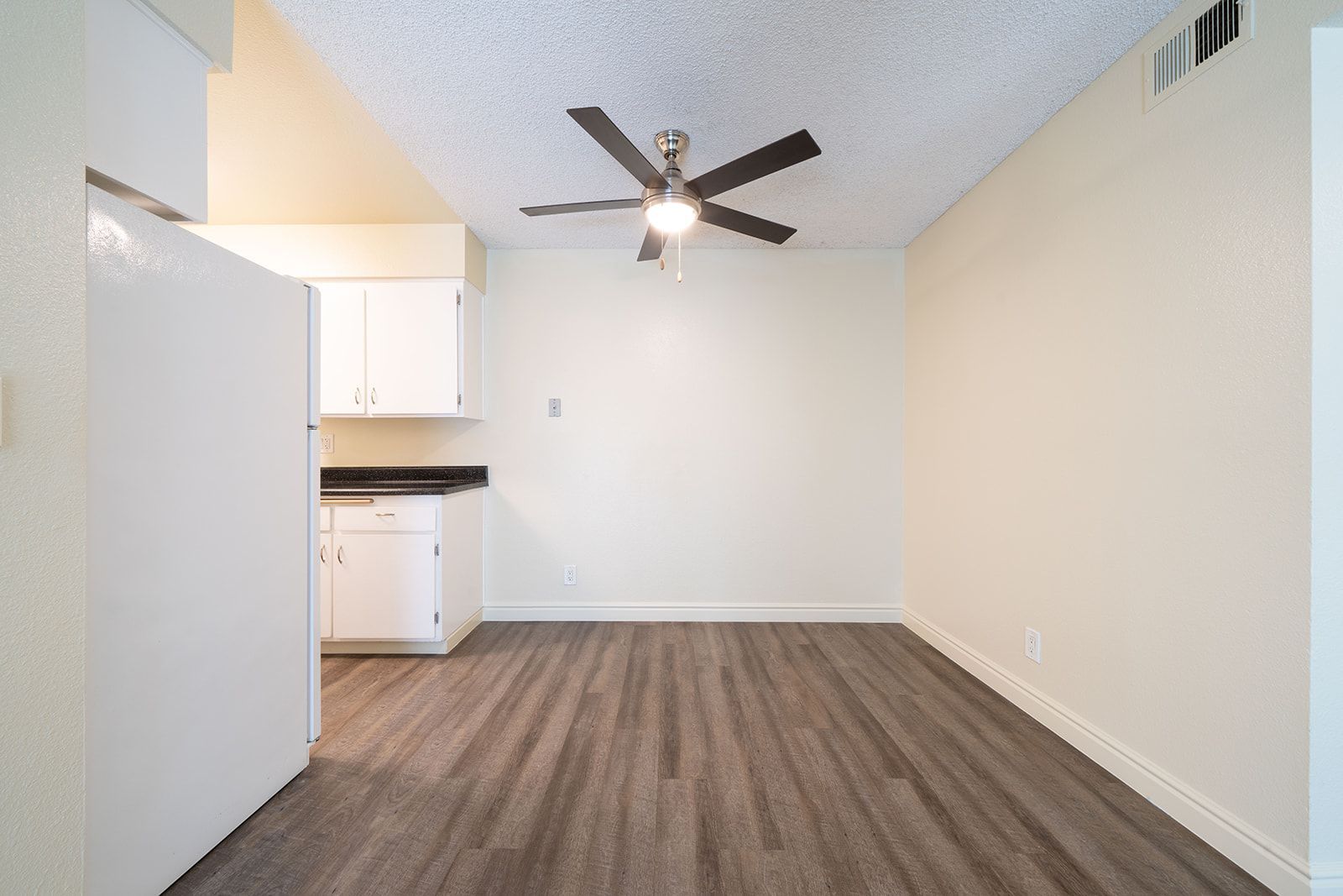 An empty room with a ceiling fan and a kitchen in the background.