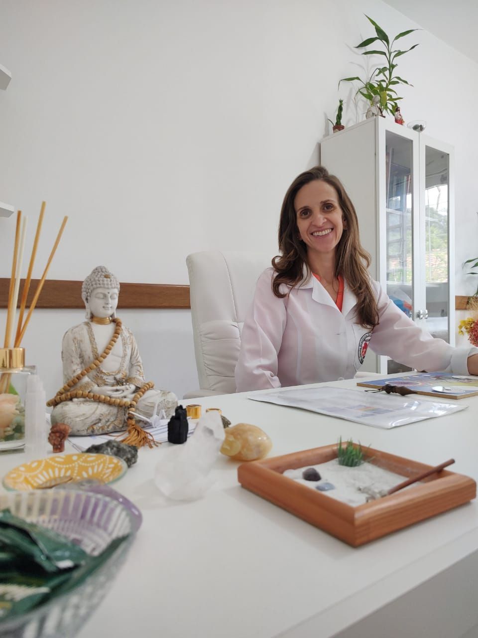 Mulher de jaleco branco sorri em frente a uma mesa com estátua de Buda, jardim zen e plantas.
