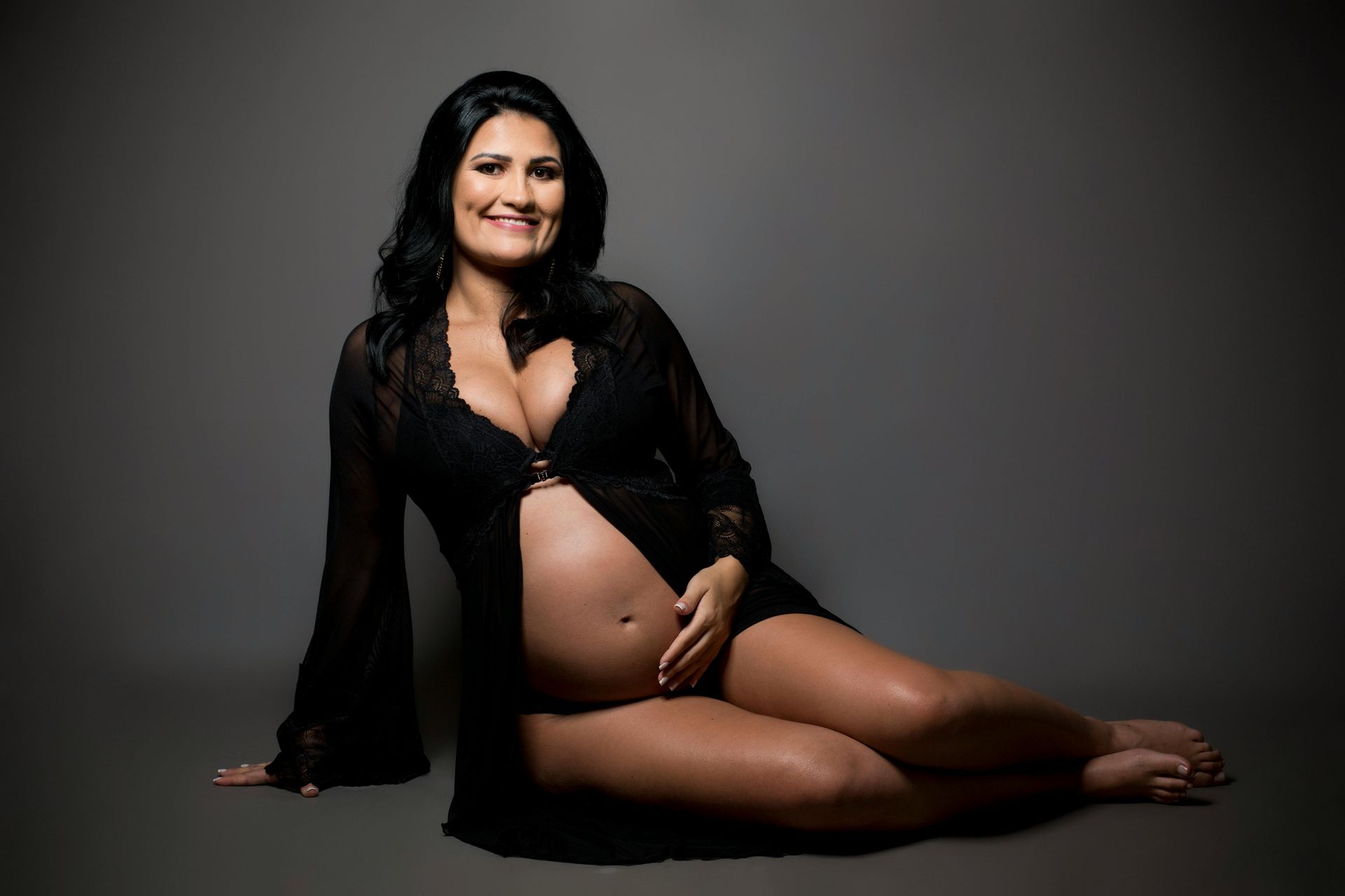 A pregnant woman in lingerie is sitting on the floor holding her belly.