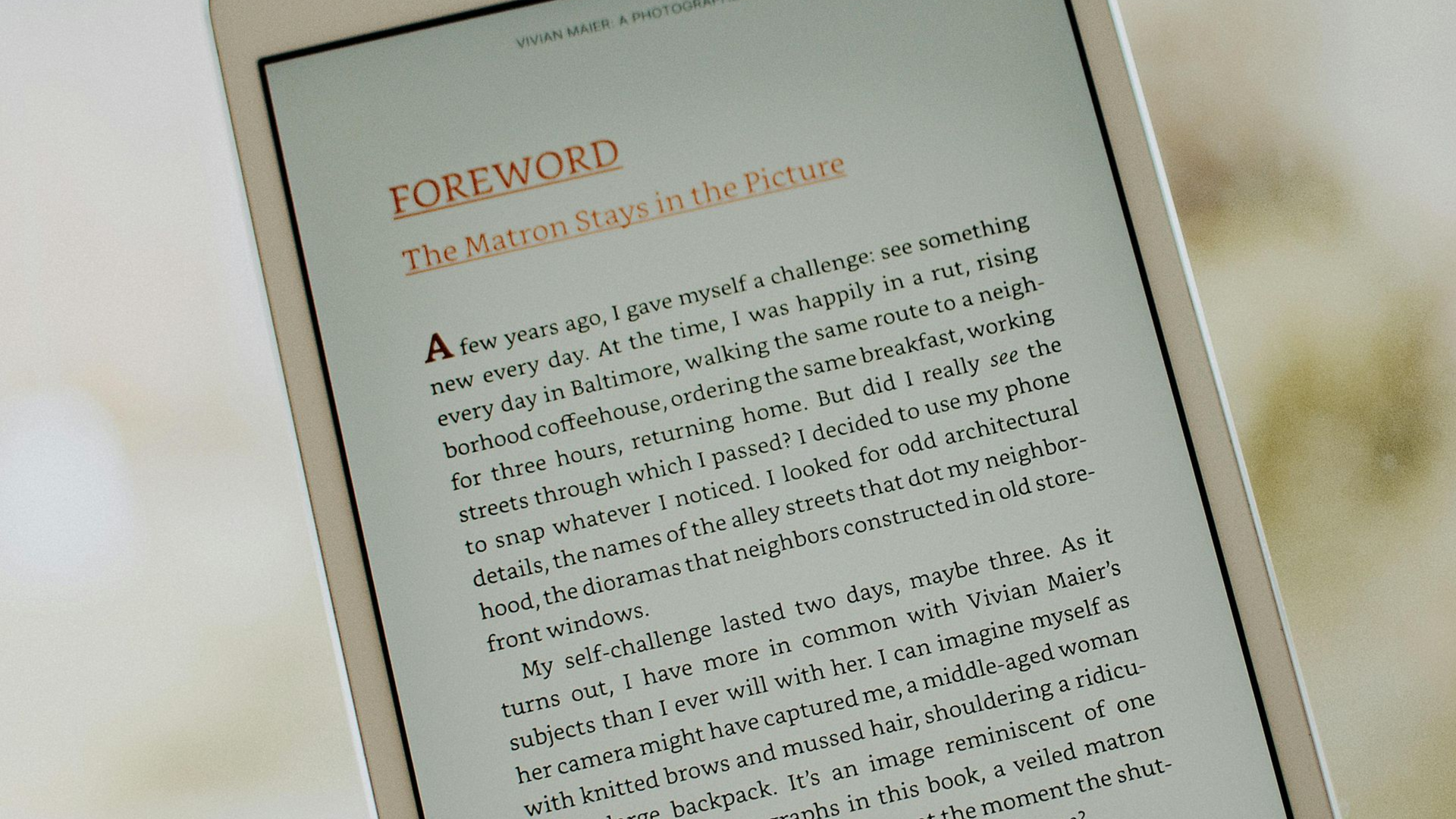 Kindle Unlimited Hand holding a white tablet displaying text outdoors. The screen shows lines of text and a red heading.