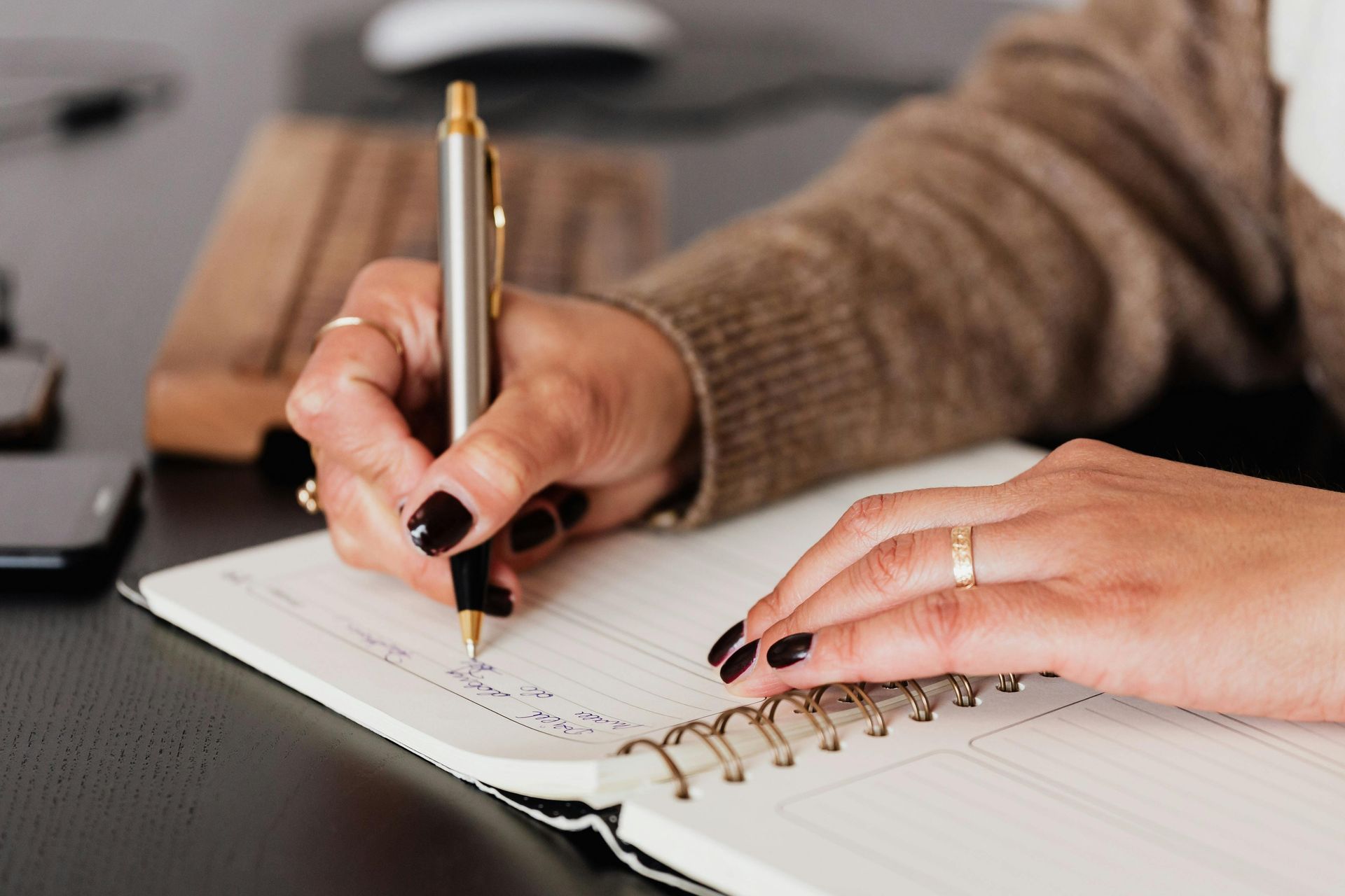 Person writing in a notebook with a silver pen, black nails, on a desk.