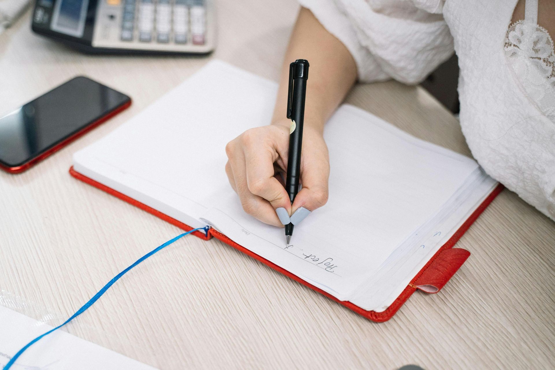 Person writing in a red notebook with a black pen, sitting at a desk. A phone and calculator are visible.