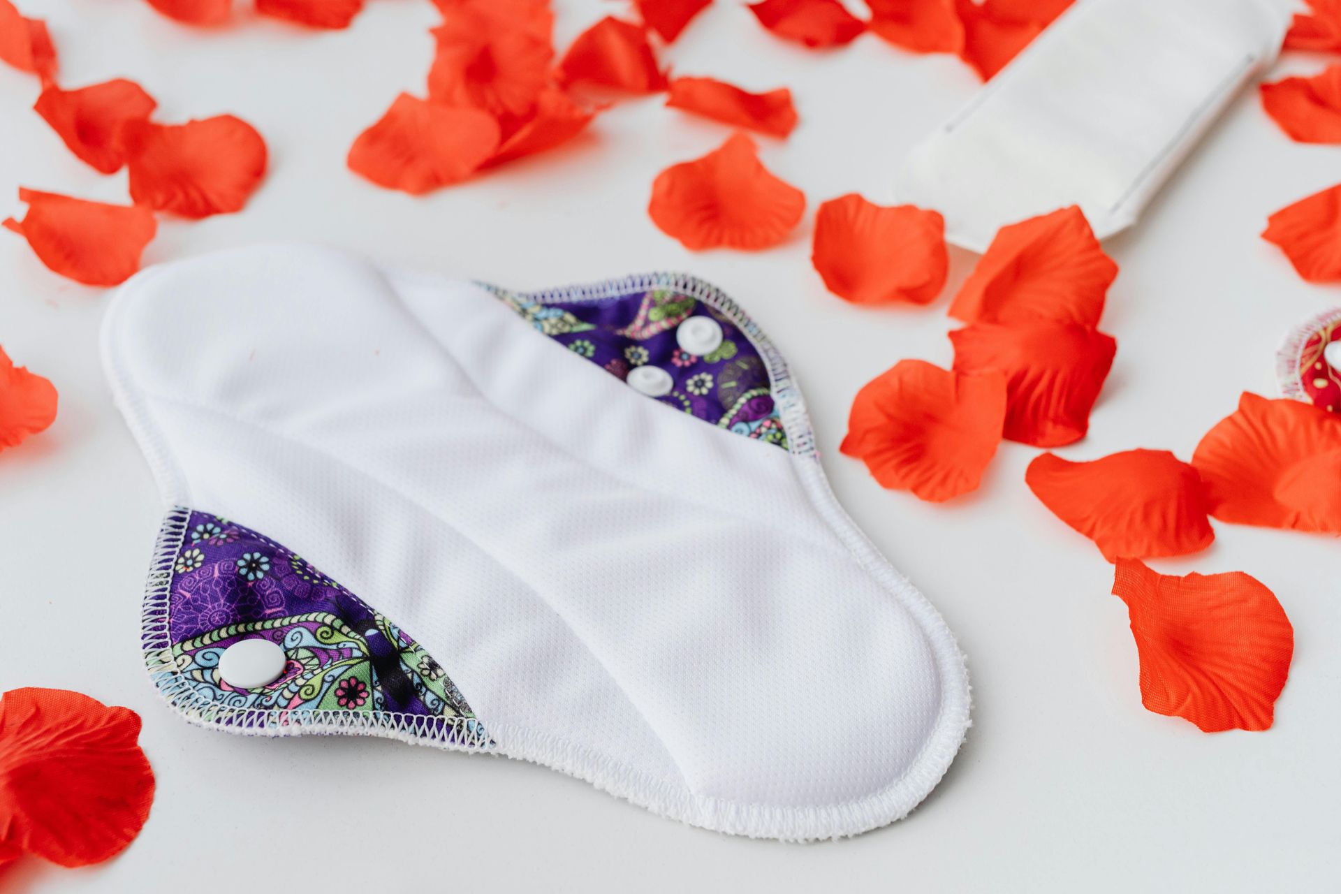 White reusable menstrual pad with purple patterned wings, surrounded by red rose petals.
