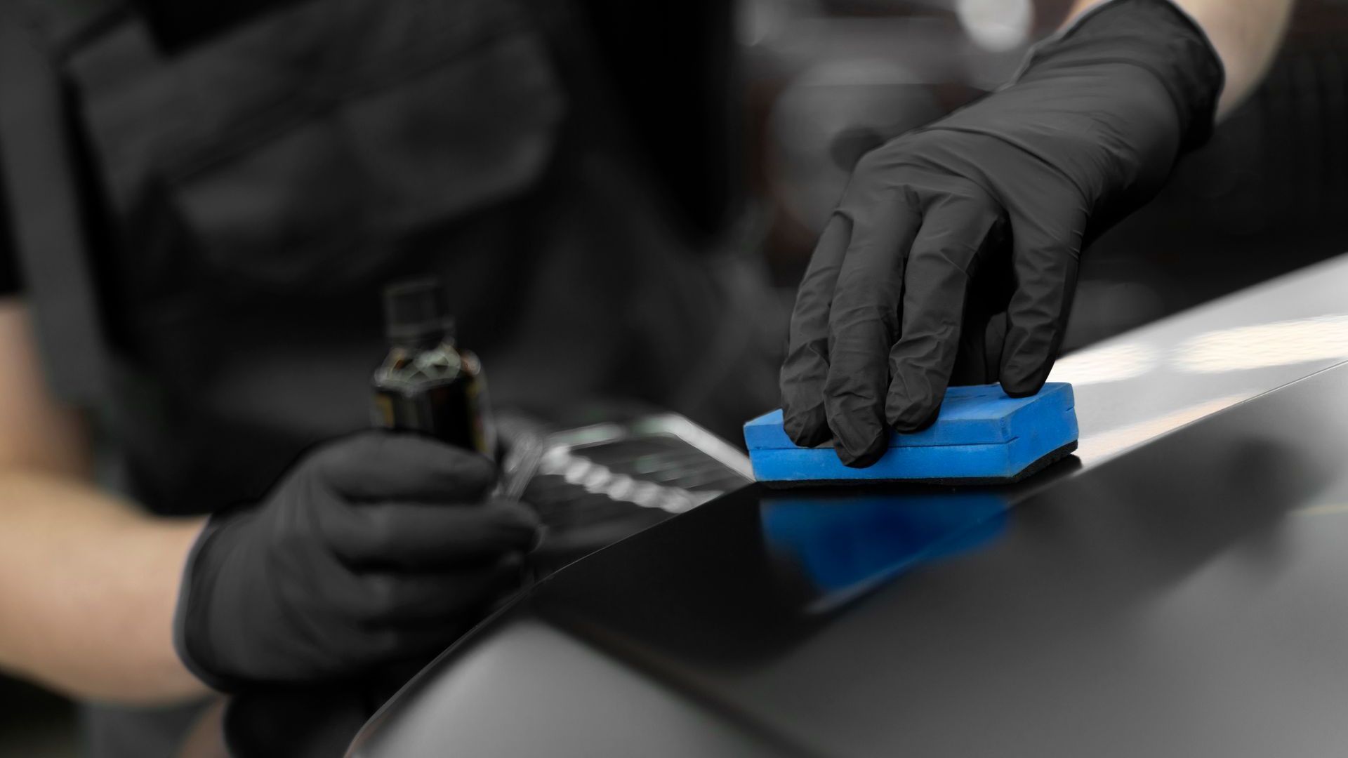 A person wearing black gloves is polishing a car with a blue sponge.