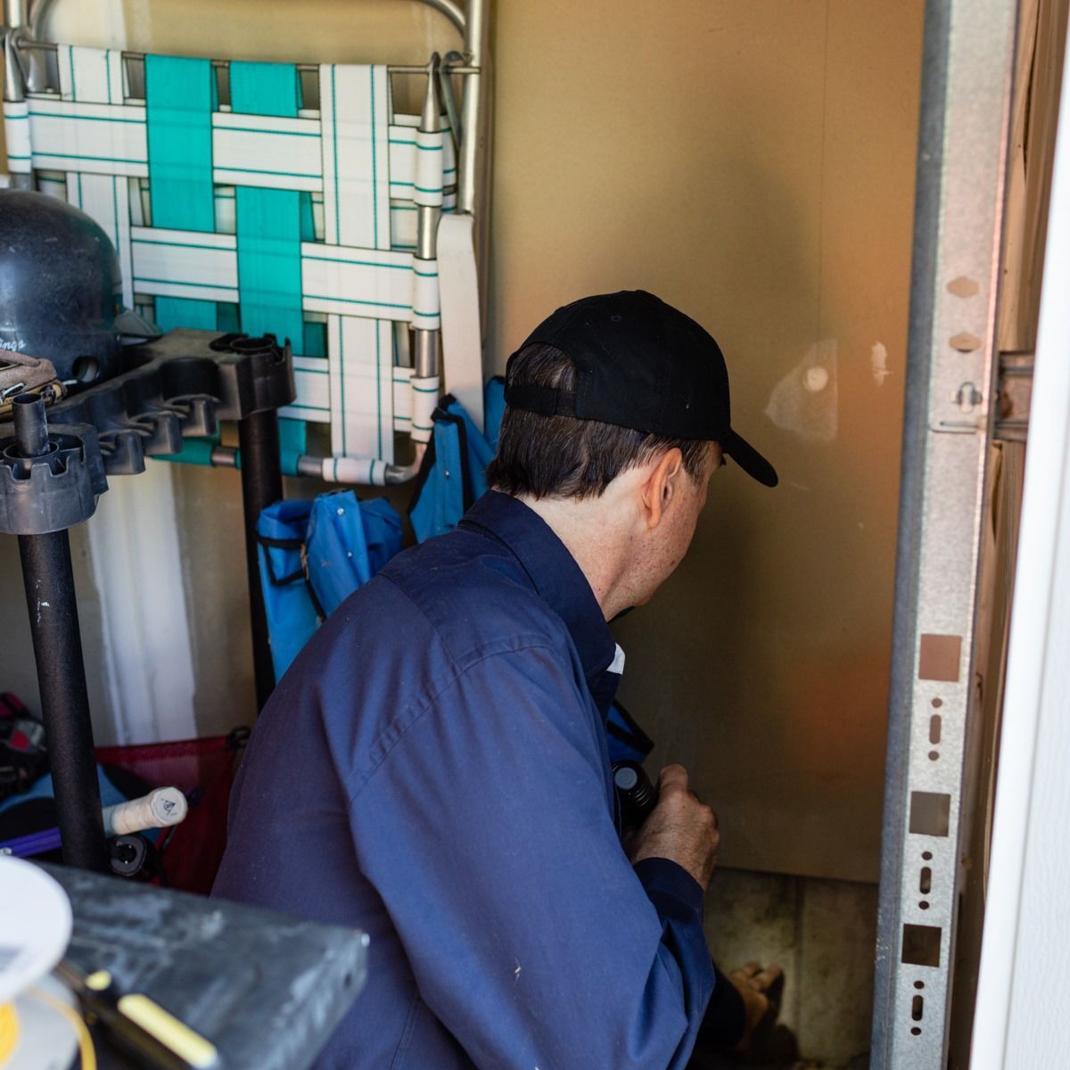 Man in blue shirt and black cap working on a garage door, near a folding chair.