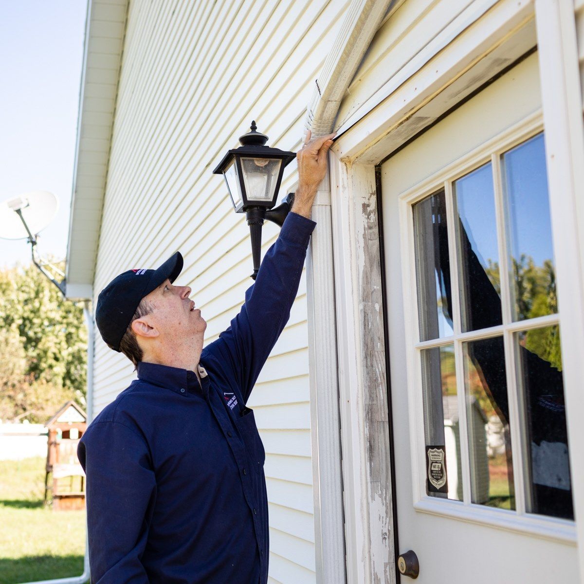 Man in blue uniform inspects door trim on a house exterior. He's wearing a cap and reaching up.