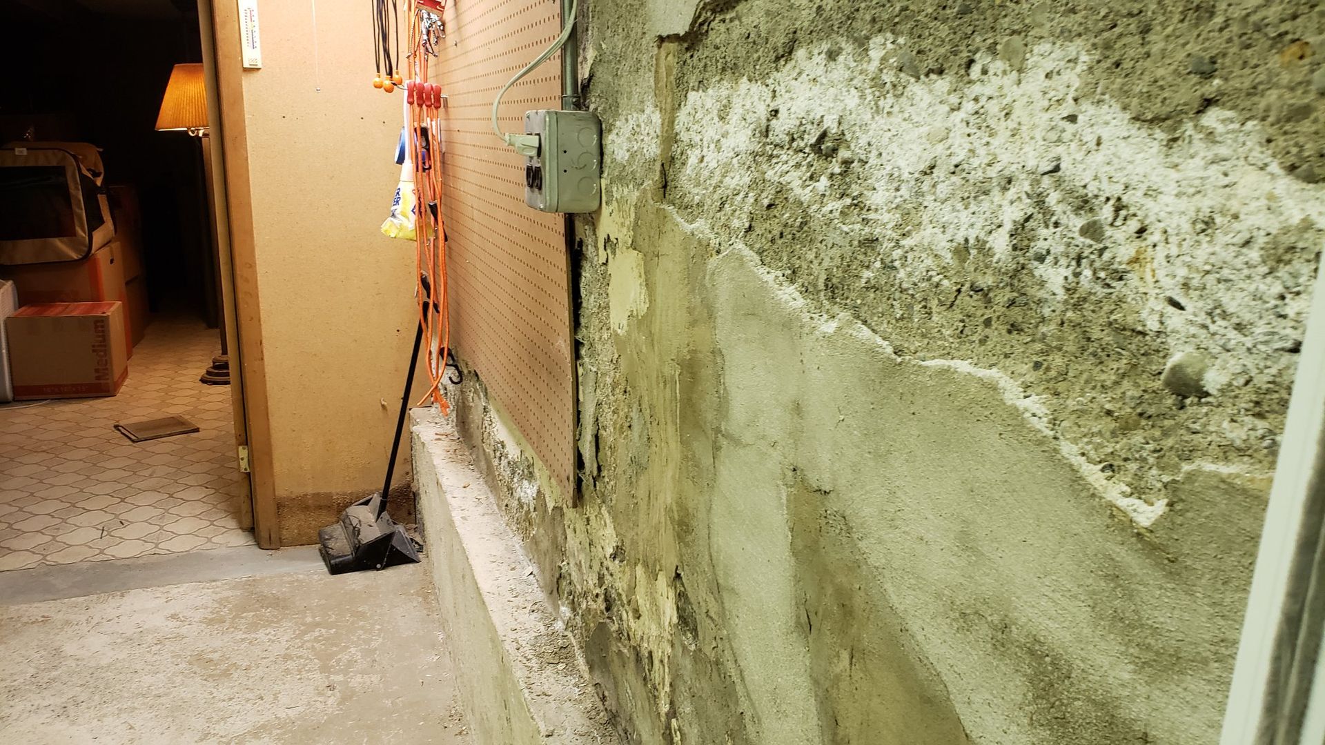 Basement with exposed concrete wall, peeling paint, electrical box, doorway to a room with a lamp.