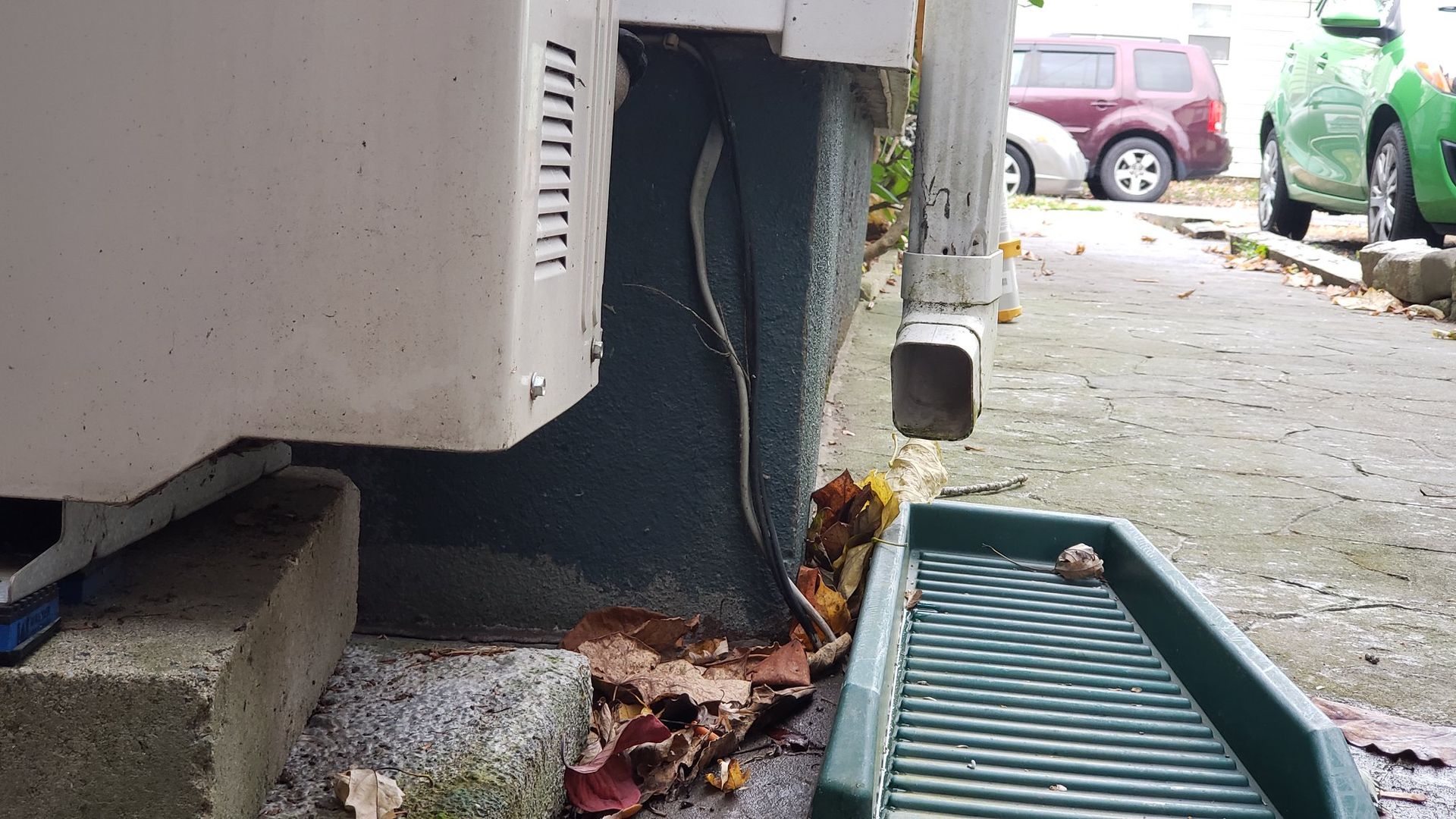 A white appliance sits on a concrete block outside. A gutter drains into a green tray, with leaves.