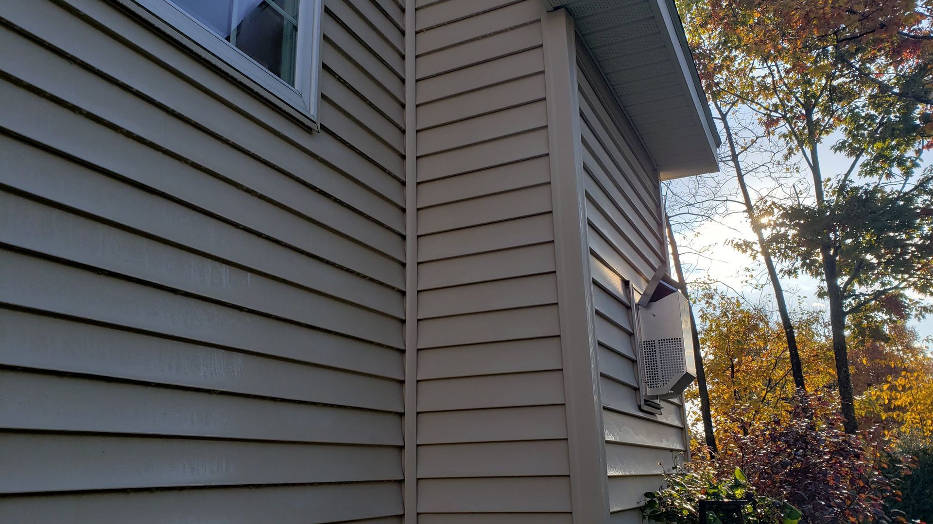Side of a beige house with horizontal siding, a white trim and window, and trees in the background.