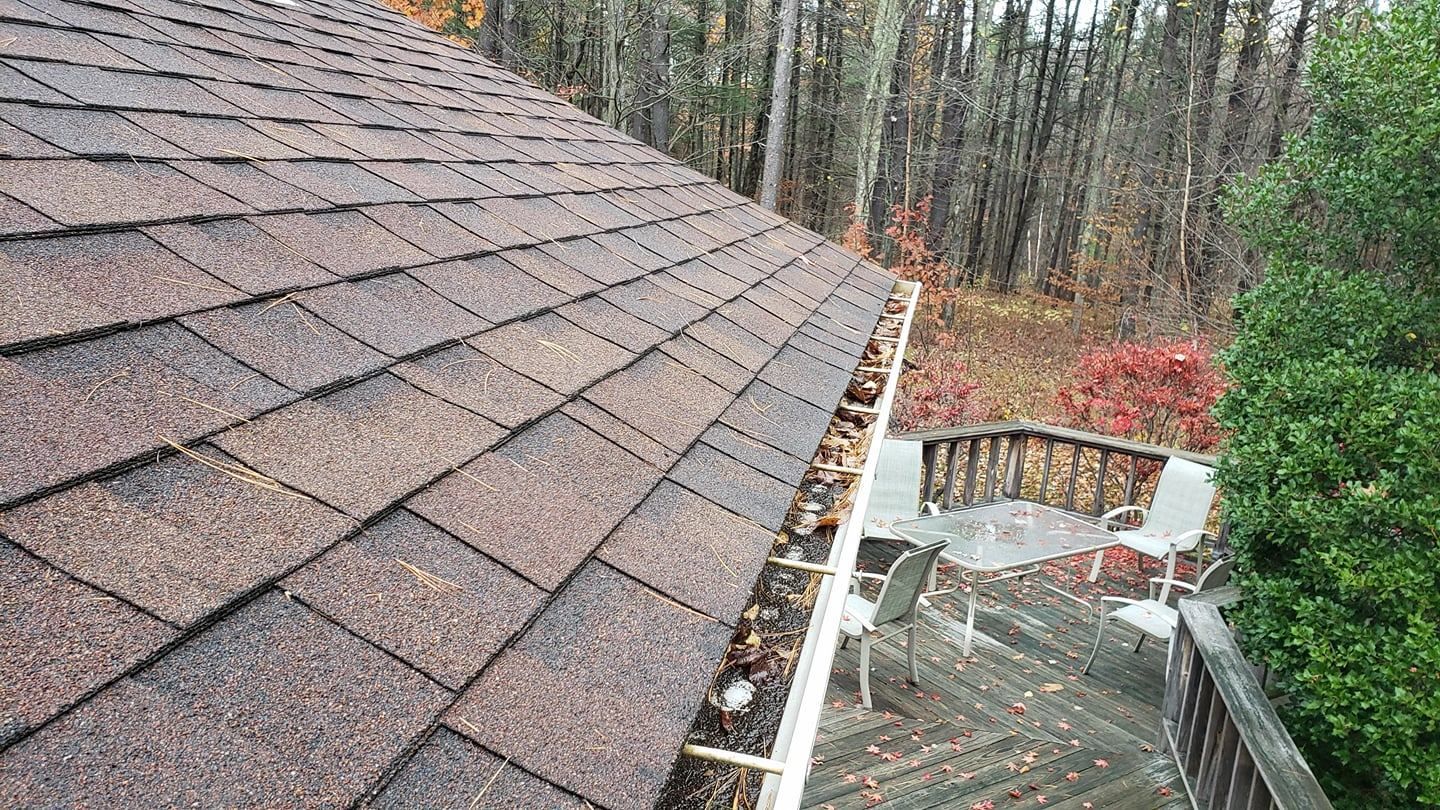 Brown shingle roof with a gutter full of debris next to a deck with patio furniture and a forest background.