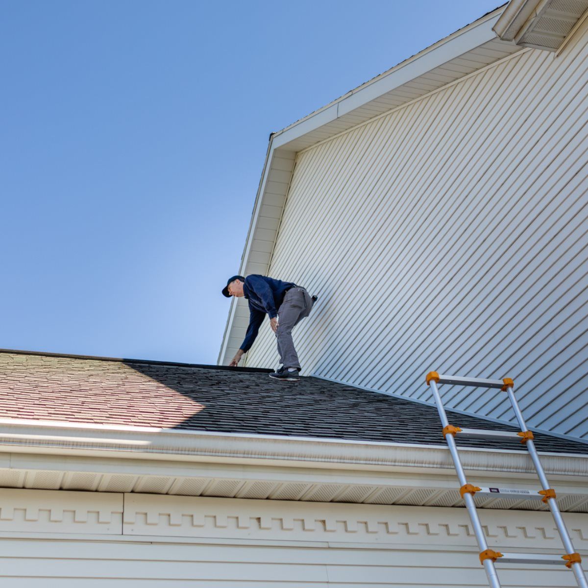 Man on a roof inspecting shingles, next to a ladder. Building is white, sky is blue.