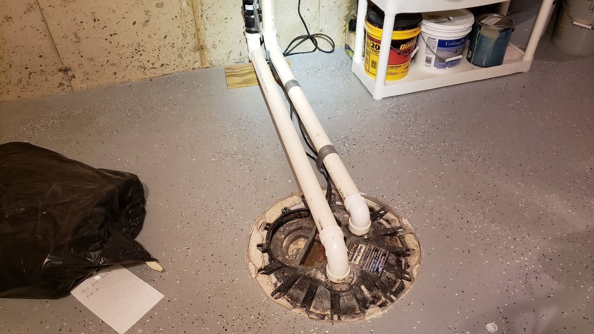 Sump pump in a basement, with pipes leading upwards. Black trash bag and white shelving unit in the background.