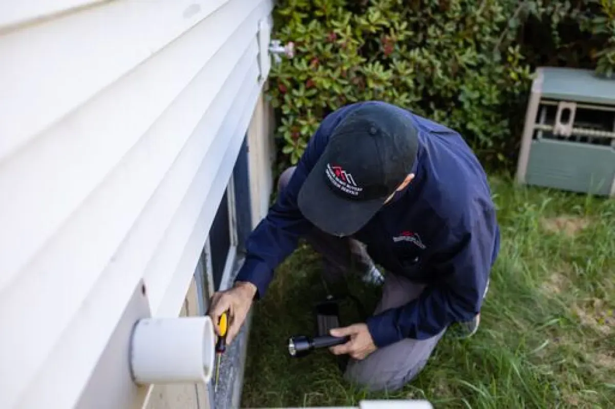 A person in work clothes examines a wall with a flashlight and utility knife, outdoors near a window.