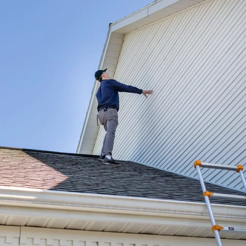 Man on roof inspecting white siding, reaching out. Blue sky, ladder present.