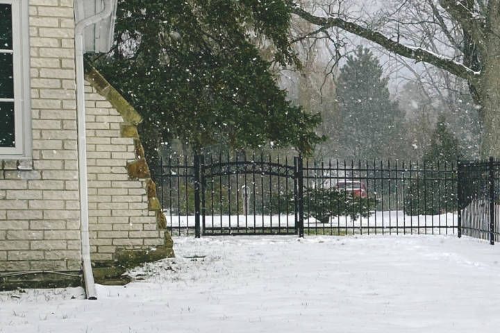 Snowy scene: a brick house, black wrought-iron fence, and snow falling on the yard.