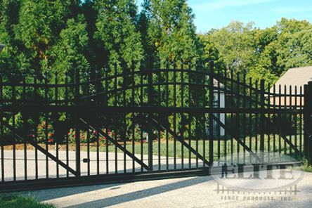 Black metal driveway gate, arched top, with vertical bars. Trees and house in the background.