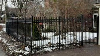 Black metal fence surrounding a yard with snow patches, bushes, and a house.