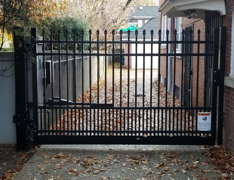 Black metal security gate, leading to a driveway with fallen leaves, set between a brick building and a concrete wall.